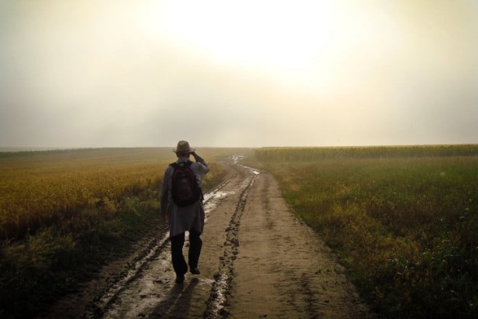 A lone traveller with a backpack walking along a quiet dirt road at sunrise
